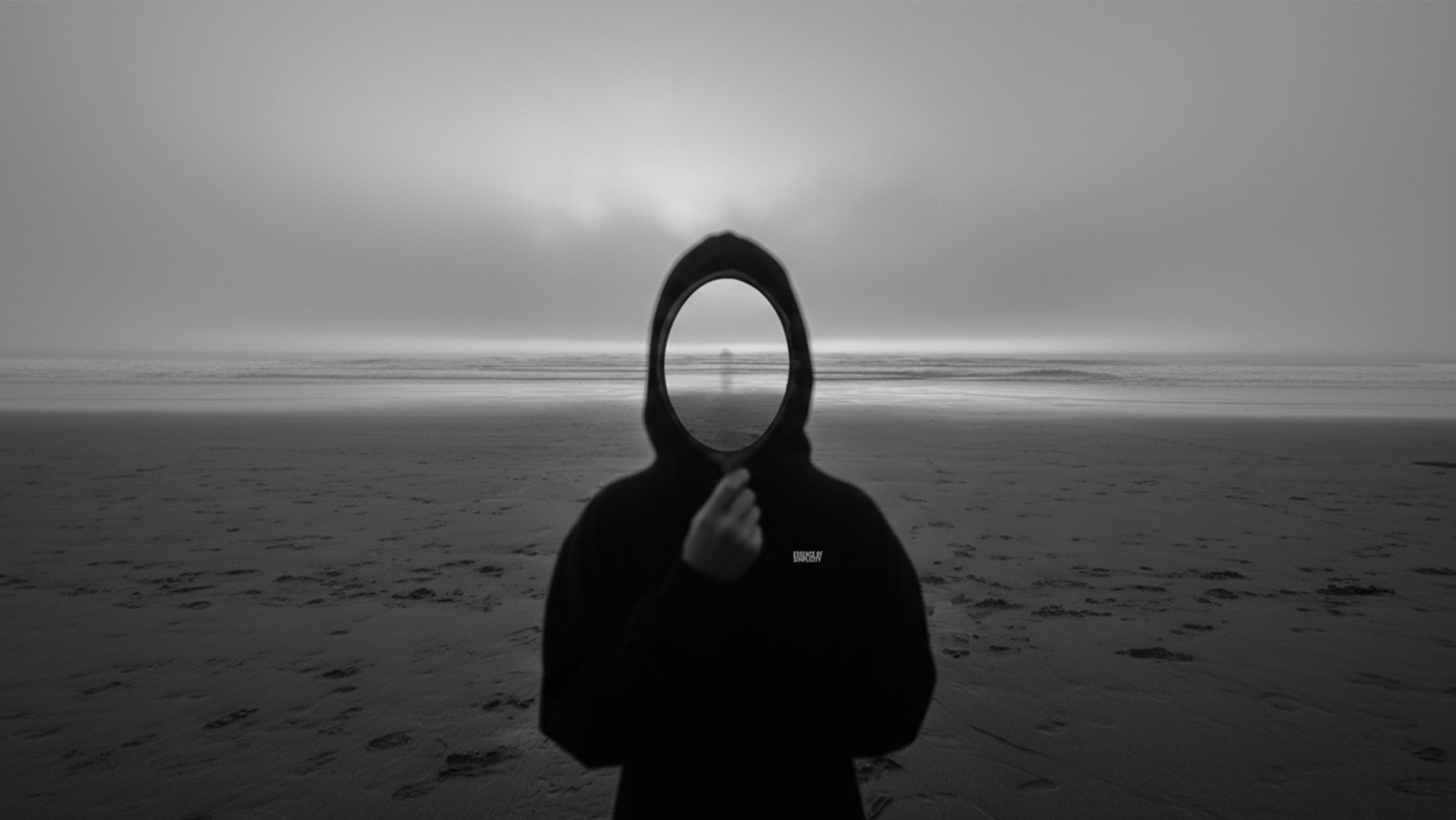 Person in a dark hooded outfit standing on a beach with a dark sky.